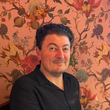 Alex Sapenuk — dark curly hair, black shirt, in front of ornate floral wallpaper